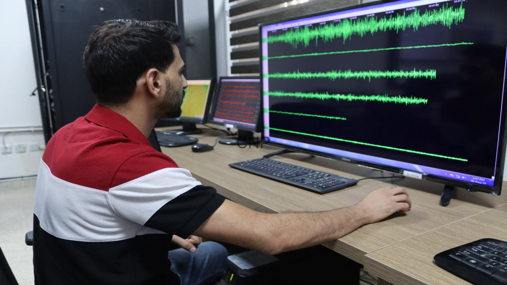 image - Center News Network Man in a red shirt sits at a desk, watching a large monitor displaying green audio waveforms.