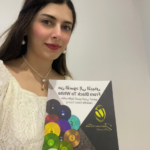 Young woman wearing a white lace top and pearl necklace holds a colorful brochure with circular color swatches and Arabic text on the cover.