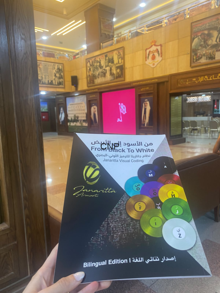 Close-up of a brochure cover titled 'From Black To White' about Janaritta Visual Coding, with colorful circular discs, held in a wood-paneled lobby entrance.