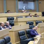 Parliamentary chamber with wooden desks and black leather chairs; several men talk in the foreground while a woman in a white hijab writes at a desk nearby.