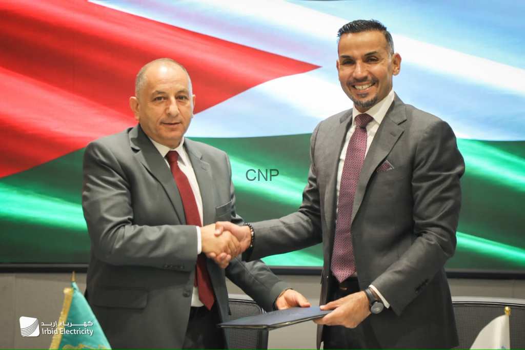 Two men in suits shake hands over a signed document, with a Jordanian flag backdrop and Irbid Electricity logo nearby, symbolizing a formal agreement.