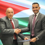 Two men in suits shake hands over a signed document, with a Jordanian flag backdrop and Irbid Electricity logo nearby, symbolizing a formal agreement.