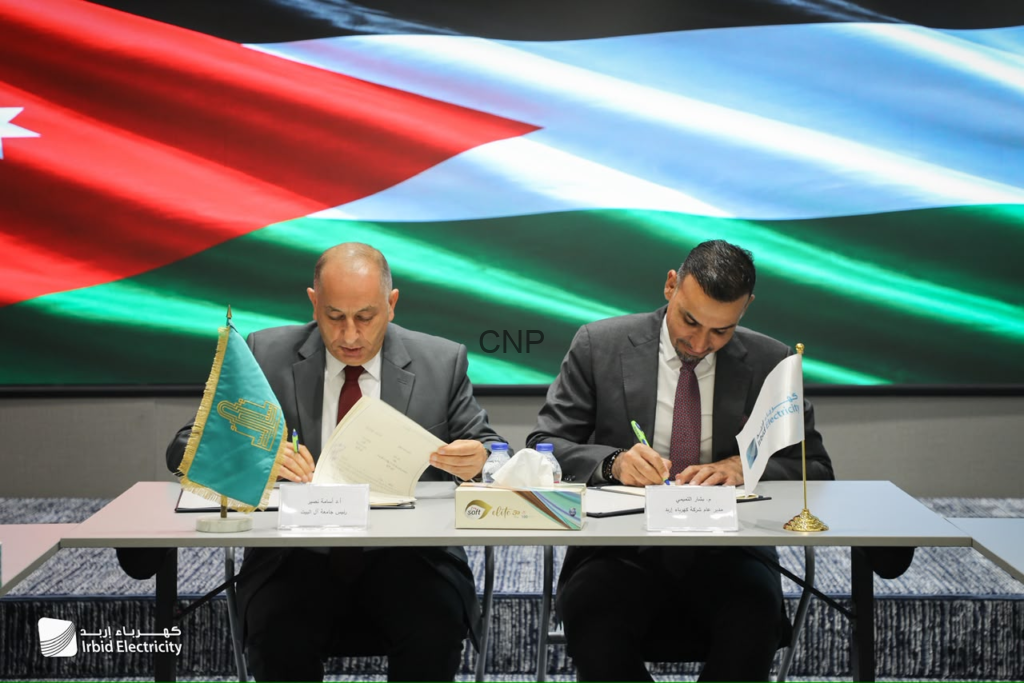 image - Center News Network Two suited men sign documents at a desk during a formal ceremony, with flags and a large flag backdrop behind them, and Irbid Electricity branding on the table.