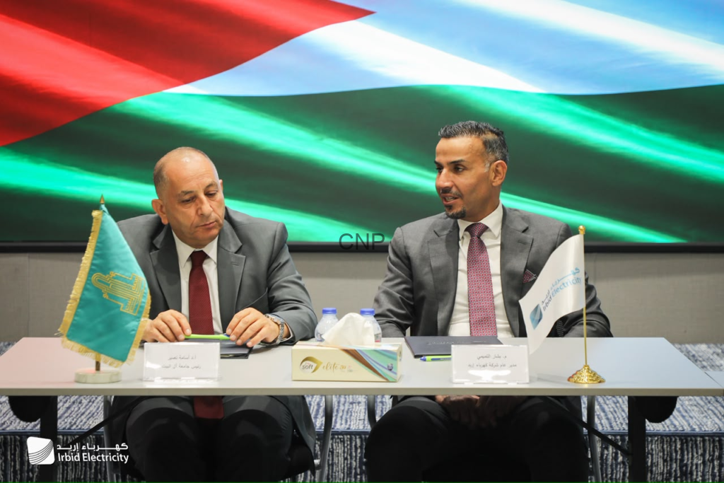 image - Center News Network Two men in suits sit at a conference table with flags, tissue box, and nameplates during a meeting, Irbid Electricity branding visible.