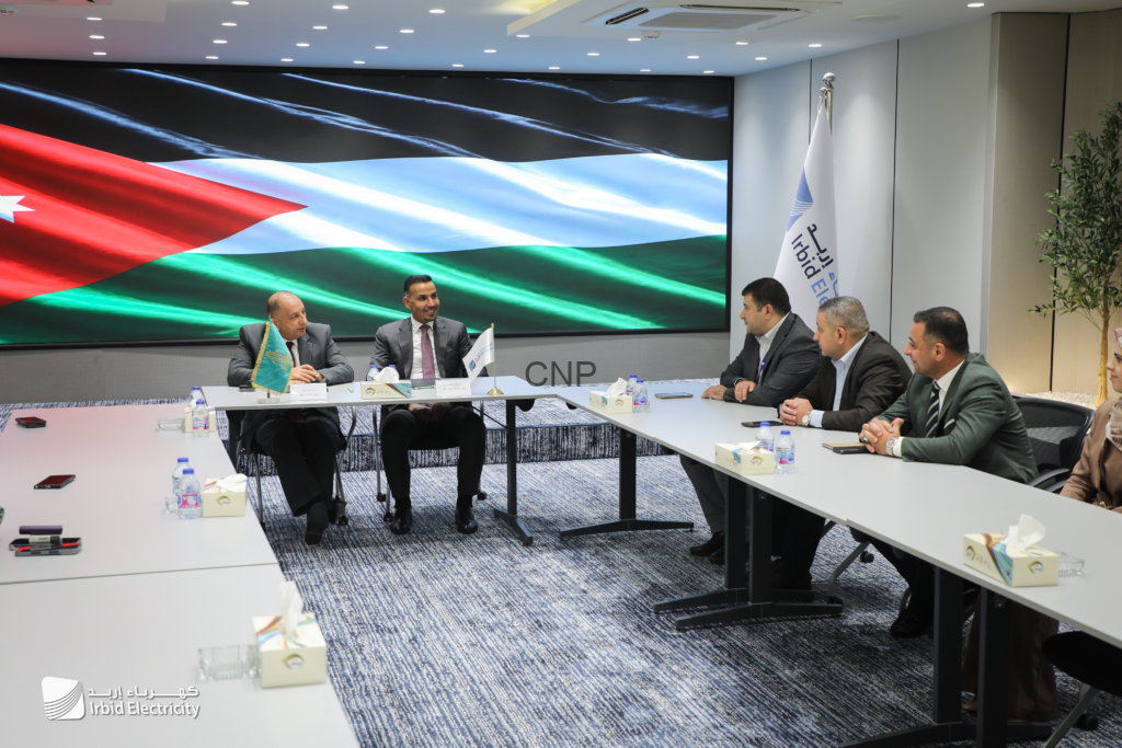 image - Center News Network Group of men in suits seated at a long conference table in a meeting room, with a large screen showing a waving Jordanian flag in the background and Irbid Electricity branding visible.