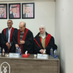 Five academics in black robes with red trim stand behind a conference table during a formal ceremony; portraits hang on the wall behind them.