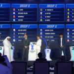Stage with a large screen showing Groups A–H for a football draw; two presenters shake hands while trophies sit on pedestals and an audience watches.