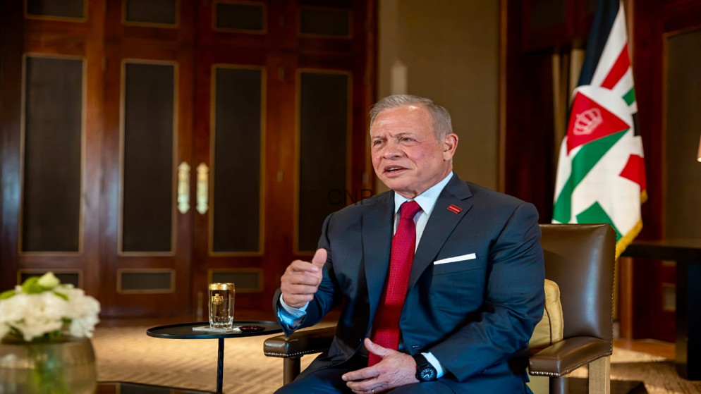 image - Center News Network Man in a dark suit and red tie sits in a leather chair, gesturing with his right hand in a formal office setting with a flag in the background, a glass of water on a small table nearby.