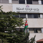 Front view of a government building with a sign reading Ministry of Local Administration and a Jordan flag in front, partially shaded by trees.