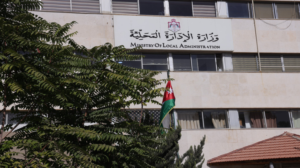 image - Center News Network Front view of a government building with a sign reading Ministry of Local Administration and a Jordan flag in front, partially shaded by trees.