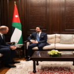 Two men in dark suits sit on cream sofas facing each other in a wood-paneled room with Jordanian flags behind them, a coffee table between them.