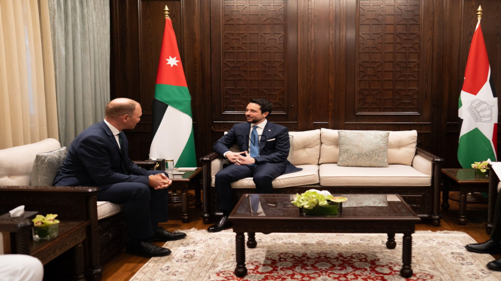 image - Center News Network Two men in dark suits sit on cream sofas facing each other in a wood-paneled room with Jordanian flags behind them, a coffee table between them.
