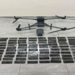 Drone with cargo bay open above a grid of numerous rectangular black packets laid out on a gray tiled floor, suggesting a seized drug case.