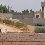 Group of people sitting under a white canopy on a dirt road beside a low stone wall, in a construction/neutral development site with a modern building in the background.