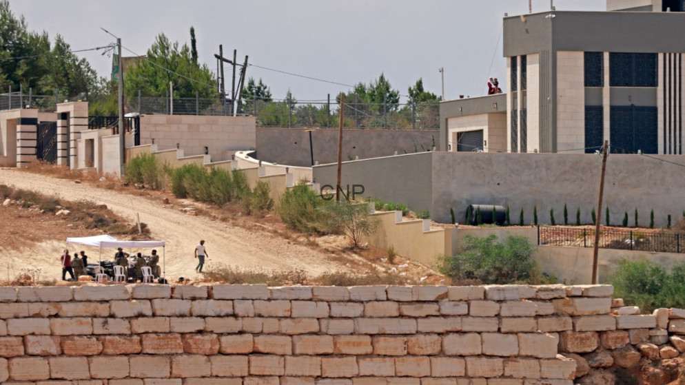 Group of people sitting under a white canopy on a dirt road beside a low stone wall, in a construction/neutral development site with a modern building in the background.
