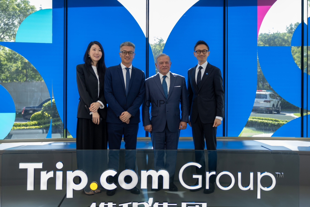 Four professionals in formal attire pose for a group photo in front of a blue glass wall with white shapes and Trip.com Group signage in the foreground.