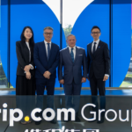 Four professionals in formal attire pose for a group photo in front of a blue glass wall with white shapes and Trip.com Group signage in the foreground.
