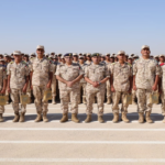 Line of soldiers in desert camouflage uniforms standing in formation on a runway, each holding a small bag, against a clear blue sky.