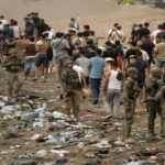Armed soldiers stand on a litter-strewn beach as a crowd of civilians forms a long line behind them.