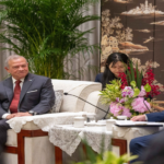 Senior man in a dark suit and red tie seated on a lace-covered sofa during a formal meeting, with others and floral arrangement nearby.