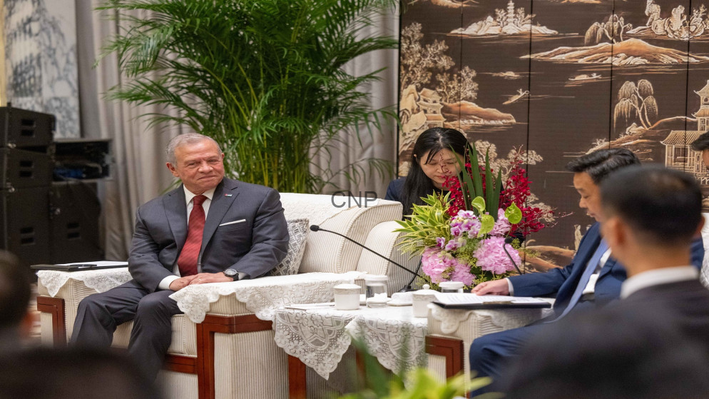 Senior man in a dark suit and red tie seated on a lace-covered sofa during a formal meeting, with others and floral arrangement nearby.