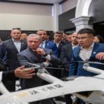 Group of professionals in suits gathered around a white quadcopter drone on display in a showroom.