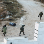 Three armed soldiers in green uniforms walk along a dirt path beside a white masonry wall with a blue drum and debris nearby on a barren landscape.