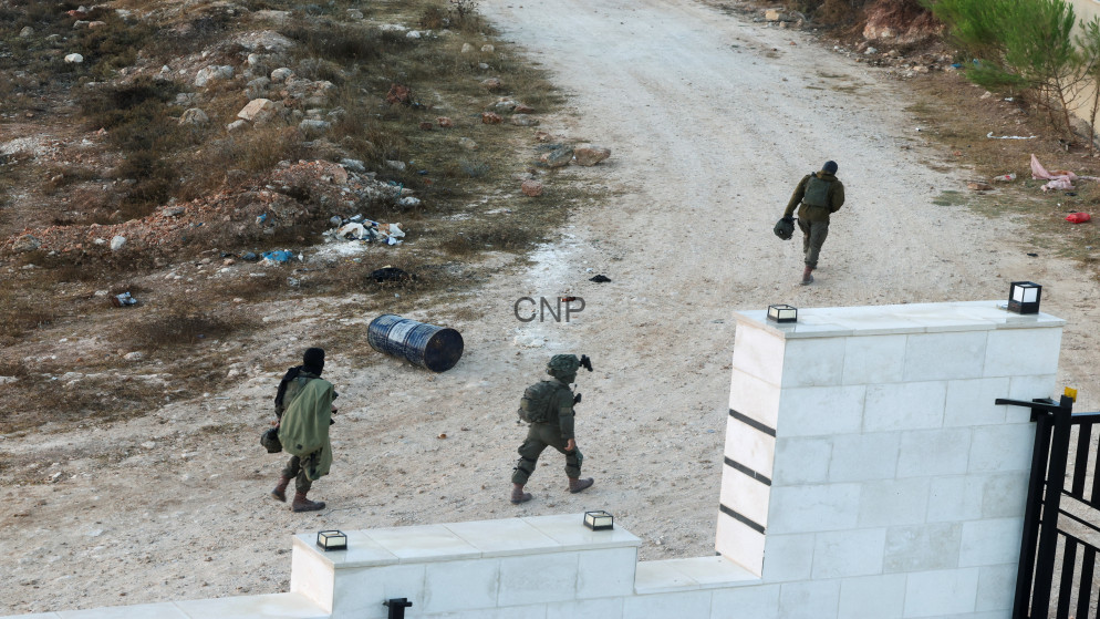Three armed soldiers in green uniforms walk along a dirt path beside a white masonry wall with a blue drum and debris nearby on a barren landscape.
