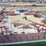 Fenced urban playground with beige shade sails, red circular posts, round seating areas, and small trees on a tiled surface; desert landscape and highway beyond.
