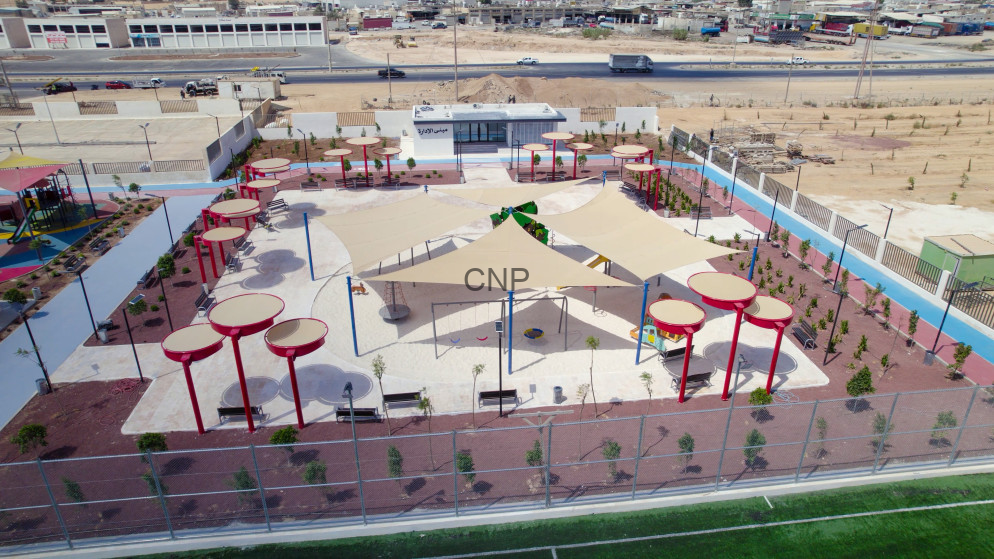 image - Center News Network Fenced urban playground with beige shade sails, red circular posts, round seating areas, and small trees on a tiled surface; desert landscape and highway beyond.