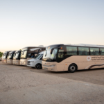 Row of beige buses parked in a dusty lot at sunset, with Arabic signage on the side.