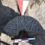 Circular, carved stone manhole cover half-buried in dirt, with a red-and-white survey marker and a 30 cm scale bar nearby.