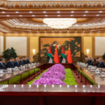 Diplomatic meeting in a grand boardroom with two long desks facing each other and flags at the center, surrounded by officials.