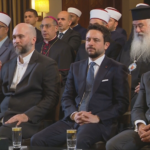 Group of men in suits and traditional religious attire seated at a formal event, facing forward.