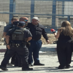 Group of police officers in tactical vests interview a man while a female reporter with a red microphone films nearby, at a fenced area