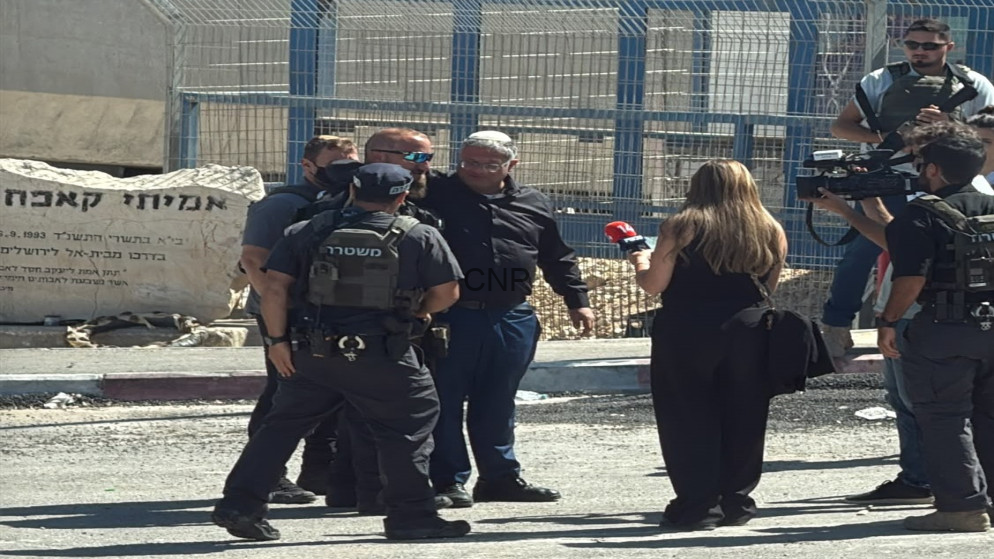 Group of police officers in tactical vests interview a man while a female reporter with a red microphone films nearby, at a fenced area