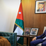 Two men sit in a formal office: a traditional-dressed Jordanian elder on a green leather sofa and a suited man on a chair, with a Jordan flag and portraits behind them.