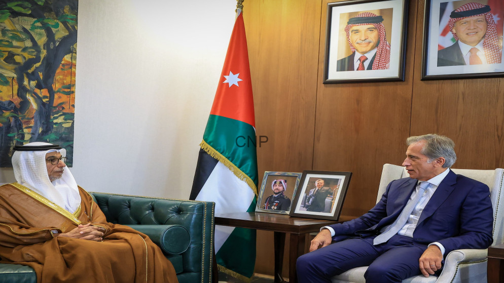 Two men sit in a formal office: a traditional-dressed Jordanian elder on a green leather sofa and a suited man on a chair, with a Jordan flag and portraits behind them.