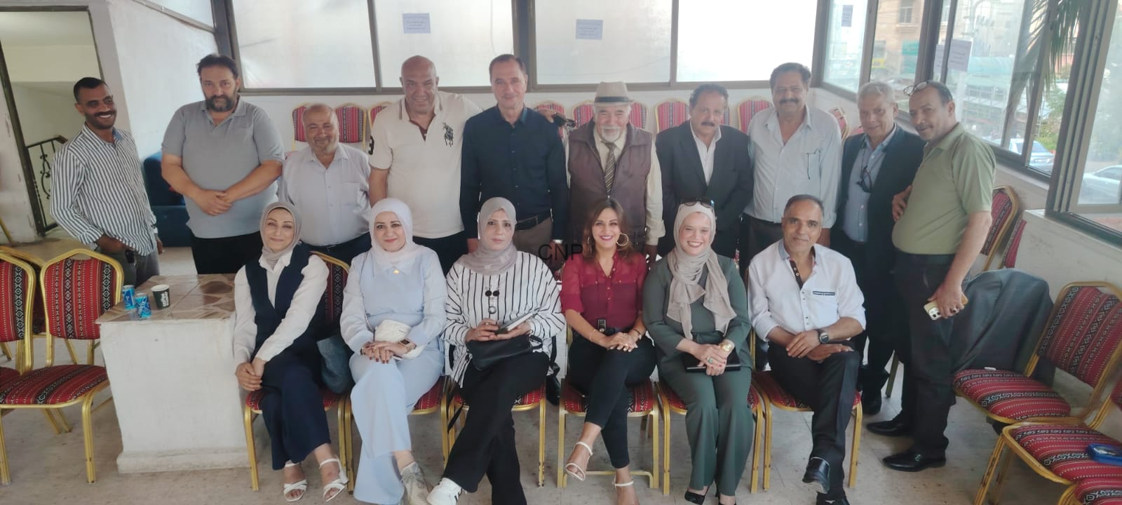 Group of 14 adults posing for a photo in a bright indoor lounge, seated in the front row and standing behind them, smiling at the camera.