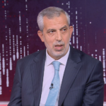 Man in a dark pinstripe suit and light blue tie seated in a TV studio during an interview, microphone clipped to the tie, purple abstract background.