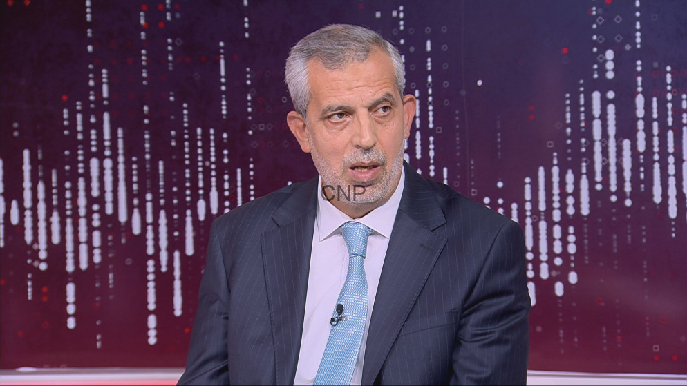 Man in a dark pinstripe suit and light blue tie seated in a TV studio during an interview, microphone clipped to the tie, purple abstract background.