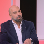 Bald man with a beard in a navy suit speaks on a pink-lit TV set.