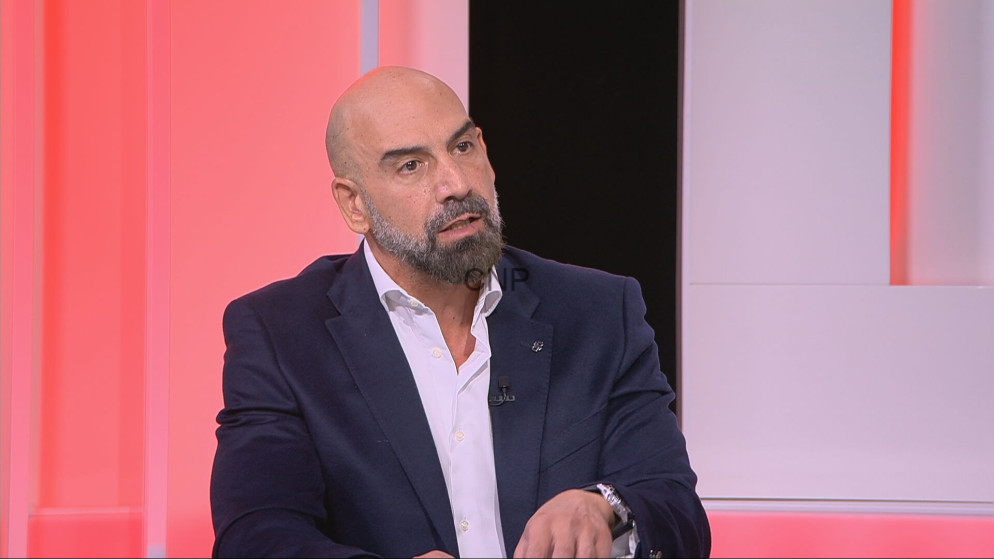 Bald man with a beard in a navy suit speaks on a pink-lit TV set.