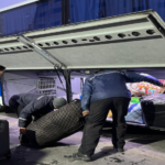 People loading suitcases and bags into the luggage compartment of a white and blue bus.