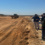 Construction site scene with a bulldozer on a dirt road as a group of workers discuss along the edge of the excavation area.