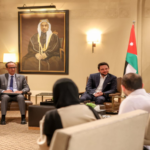 Diplomatic meeting in a formal lounge with attendees seated in chairs around coffee tables and a Jordan flag in the background