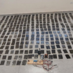 Room with dozens of small black rectangular packages laid out in neat rows on a tiled floor; foreground has cables and testing equipment.