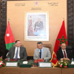 Formal meeting at a conference table with Moroccan and Jordanian flags and a framed portrait behind the speakers.