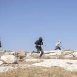 Three people run across a rocky, arid landscape under a clear blue sky, with a town in the distance.