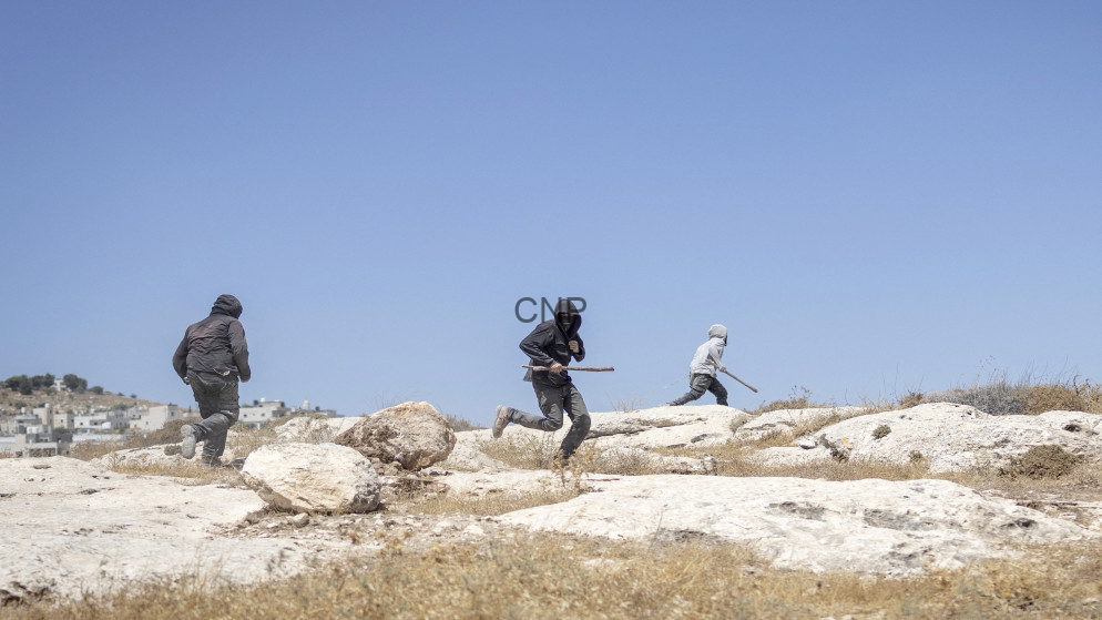 Three people run across a rocky, arid landscape under a clear blue sky, with a town in the distance.